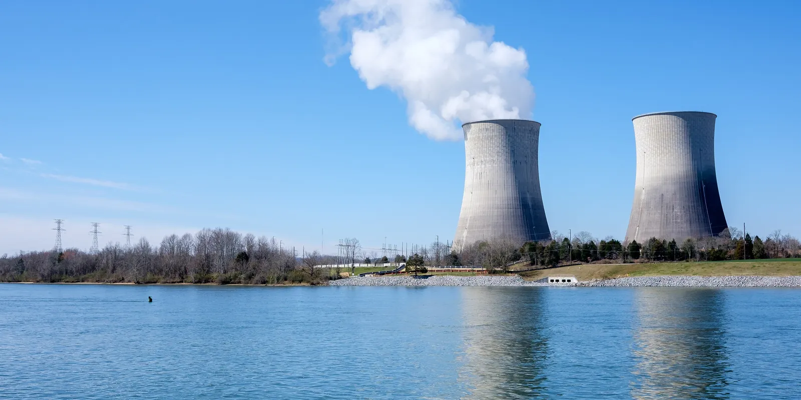 Two concrete nuclear plant cooling towers beside a lake, one releasing steam